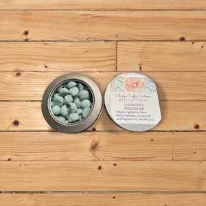 May include: A silver tin containing light blue lotion dots, next to a lid with floral design and text: "LOTION DOTS ACQUA DI GIO". The ingredients include shea butter, beeswax, and coconut oil. Net weight is 2 oz.