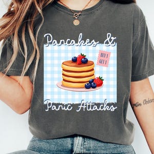 Peut inclure: T-shirt gris foncé avec un motif de crêpes garnies de myrtilles et de fraises sur une assiette rose, sur fond à carreaux bleus et blancs. Le texte indique "Pancakes & Panic Attacks". Un autocollant "Achetez-en 1, obtenez-en 1" est également visible.