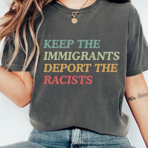 May include: Dark gray t-shirt with the text "KEEP THE IMMIGRANTS DEPORT THE RACISTS" in green, light blue, yellow, and red. The shirt is a casual style.