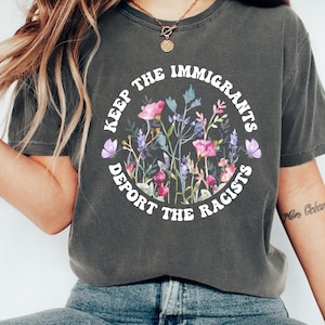 以下が含まれることがあります： 花柄のデザインと「KEEP THE IMMIGRANTS DEPORT THE RACISTS」の文字が円形に配置された濃いグレーのTシャツ。 花柄のデザインには、ピンク、紫、緑の花と2匹の紫色の蝶が含まれています。