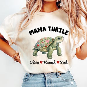 May include: Cream-colored t-shirt featuring a green turtle illustration with a floral and heart-shaped design on its shell. The text "MAMA TURTLE" is above the turtle, with names "Olivia", "Hannah", and "Jack" below.