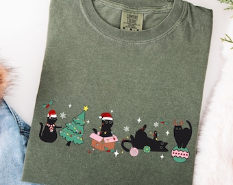 Puede incluir: Camiseta verde oliva con un diseño navideño. Presenta gatos negros con gorros de Papá Noel decorando un árbol de Navidad. Los gatos están en varias poses, con copos de nieve y adornos. La camiseta tiene cuello redondo y mangas cortas.