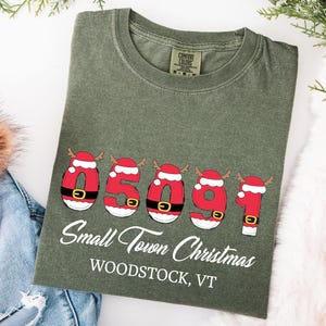 Może przedstawiać: Zielona koszulka z motywem świątecznym. Projekt przedstawia kod pocztowy "05091" w kolorze czerwonym, liczby w stylu Świętego Mikołaja oraz napis "Small Town Christmas Woodstock, VT". Koszulka jest ozdobiona świąteczną zielenią.