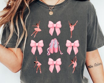 May include: A dark gray t-shirt with a graphic design featuring pink bows, ballet shoes, and black silhouettes of dancers. The text "More Ocean" is written on the right sleeve.