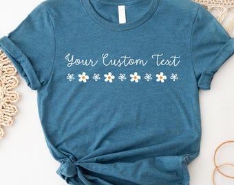 Op de afbeelding: Een tealkleurig T-shirt met opgerolde mouwen. Het shirt heeft de tekst "Your Custom Text" in een cursief lettertype, boven een rij witte madeliefjes met gele centra. Het T-shirt is aan de onderkant geknoopt.