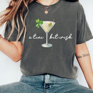 Könnte beinhalten: Dunkelgraues T-Shirt mit einem Martini-Glas-Motiv und dem Text "a tini bit irish". Das Martini-Glas hat eine grüne Olive und ein vierblättriges Kleeblatt. Das T-Shirt ist ein lässiger Stil.