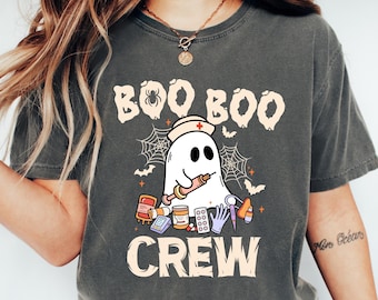 May include: Dark grey t-shirt with a Halloween design. It shows a ghost in a nurse's hat holding a syringe, with cobwebs, bats, and the words "BOO BOO CREW". The text is in a playful font, perfect for the spooky season.