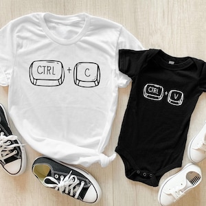 以下が含まれることがあります： 黒い文字とグラフィックが描かれた白いTシャツ2枚。大きい方のTシャツには「CTRL + C」の文字が、小さい方のTシャツには「CTRL + V」の文字が書かれています。