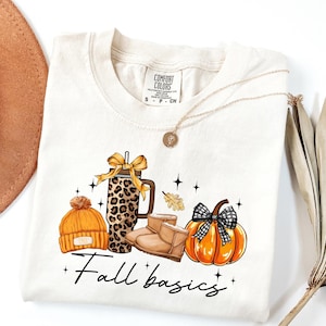 May include: Cream-colored t-shirt with a graphic of fall-themed items: an orange beanie, a leopard-print tumbler, brown boots, and a pumpkin with a black and white bow. The text "Fall basics" is written in cursive.