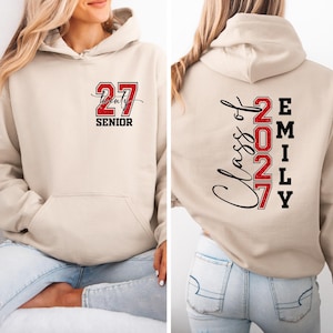 May include: A beige hooded sweatshirt featuring a red glittery number 27 and the word SENIOR on the front. The back displays the text Class of 2027 in black script and red glittery numbers, alongside the name EMILY printed vertically in black block letters.