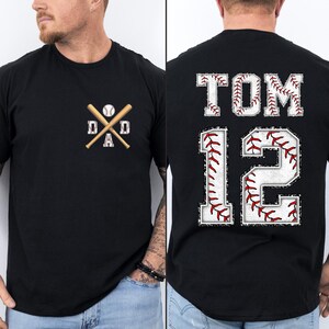 May include: Black t-shirt with a baseball-themed design. The front features crossed baseball bats, a baseball, and the word "DAD." The back displays "TOM 12" in a baseball stitch font.