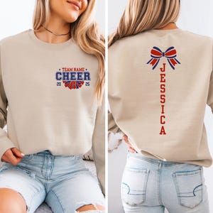 May include: A beige sweatshirt with a cheerleading design on the front and a name on the back. The front design includes the words "Team Name Cheer" with a pom-pom graphic and the numbers "20" and "25". The back design features a red and blue bow and the name "Jessica" written vertically.