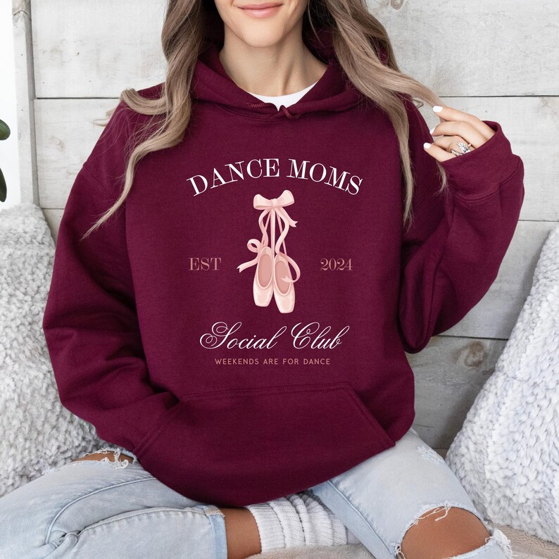 May include: Burgundy hoodie with a white graphic that says "Dance Moms Est 2021 Social Club Weekends Are For Dance" with a pair of pink ballet shoes.