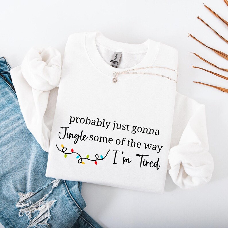 May include: White sweatshirt with the text "probably just gonna Jingle some of the way I'm Tired" in black script. A string of colorful Christmas lights is below the text. A silver necklace with a small pendant is on the sweatshirt.