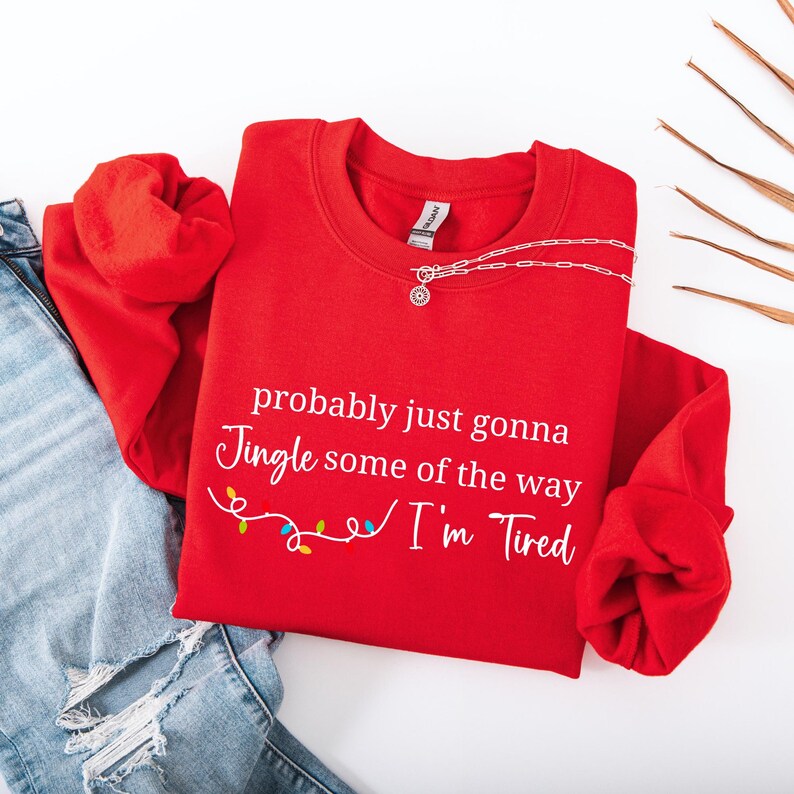 May include: A bright red sweatshirt with the text "probably just gonna Jingle some of the way I'm Tired" in white script. A silver necklace with a pendant is on the sweatshirt. The sweatshirt is folded next to a pair of blue jeans.