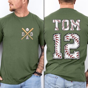 May include: Olive green baseball-themed t-shirt. The front features a crossed baseball bat and ball design with the word "DAD". The back displays the name "TOM" and the number "12", both with baseball stitching.
