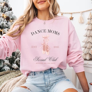 May include: Pink sweatshirt with the text "Dance Moms Social Club Est 2025 Weekends are for Dance" and a pair of pink ballet shoes.