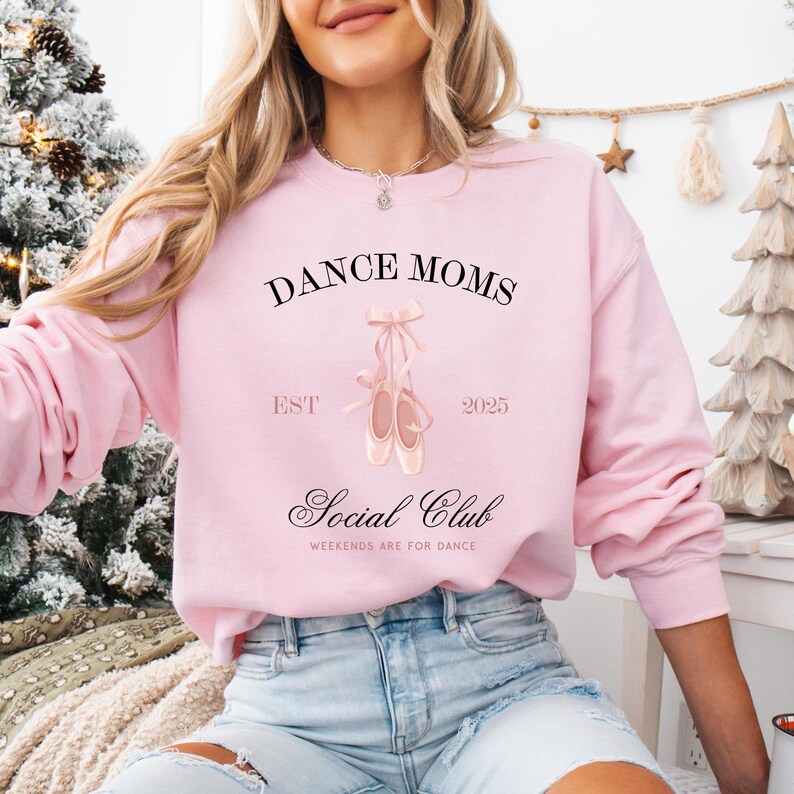 May include: Pink sweatshirt with the text "Dance Moms Social Club Est 2025 Weekends are for Dance" and a pair of pink ballet shoes.