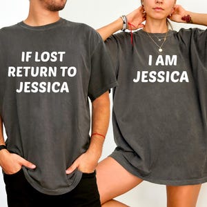 Könnte beinhalten: Zwei anthrazitfarbene T-Shirts mit weißem Text. Auf einem Shirt steht "IF LOST RETURN TO JESSICA", auf dem anderen "I AM JESSICA". Die Shirts haben eine lockere Passform.