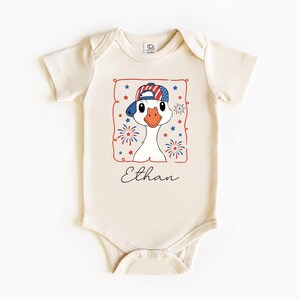 May include: Cream-colored baby onesie featuring a cartoon goose wearing a patriotic baseball cap with red, white, and blue stars and stripes. The design includes fireworks and stars, with the name "Ethan" in script.