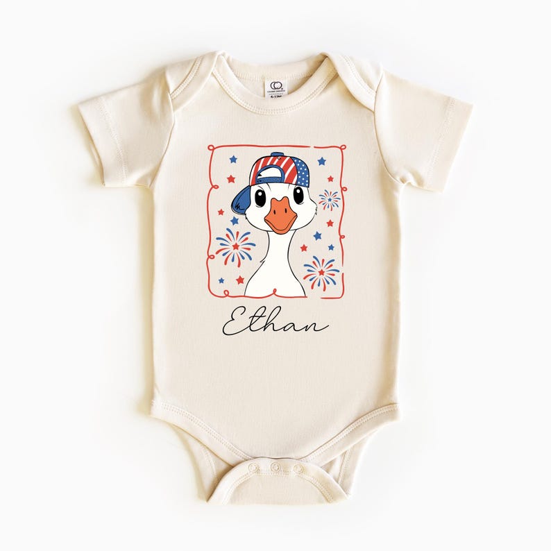 May include: Cream-colored baby onesie featuring a cartoon goose wearing a patriotic baseball cap with red, white, and blue stars and stripes. The design includes fireworks and stars, with the name "Ethan" in script.