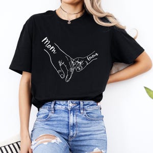 Puede incluir: Camiseta negra con un contorno blanco de dos manos que se sostienen. El texto "Mom" est&aacute; escrito en una mano y "Emma" en la otra.