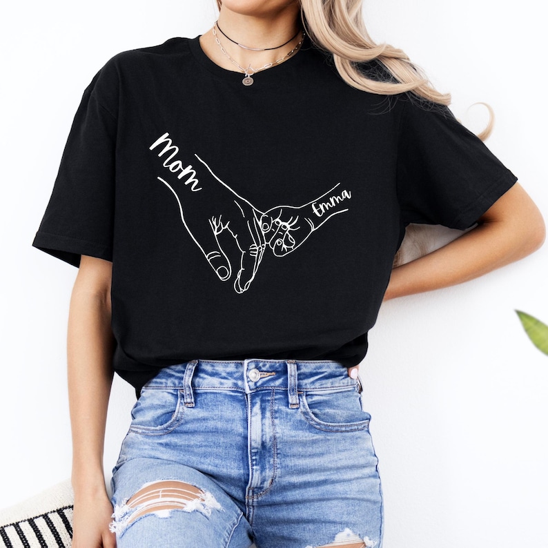 Puede incluir: Camiseta negra con un contorno blanco de dos manos que se sostienen. El texto "Mom" est&aacute; escrito en una mano y "Emma" en la otra.