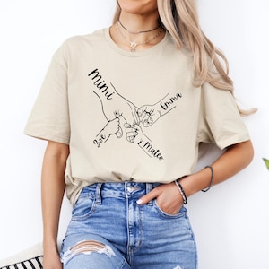 Puede incluir: Una camiseta beige con un gr&aacute;fico de cuatro manos que se agarran. Los nombres "Mimi", "Emma", "Zoe" y "Mateo" est&aacute;n escritos en las manos.