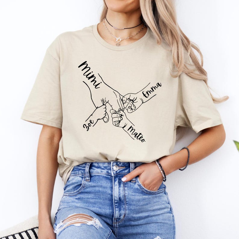 Puede incluir: Una camiseta beige con un gr&aacute;fico de cuatro manos que se agarran. Los nombres "Mimi", "Emma", "Zoe" y "Mateo" est&aacute;n escritos en las manos.
