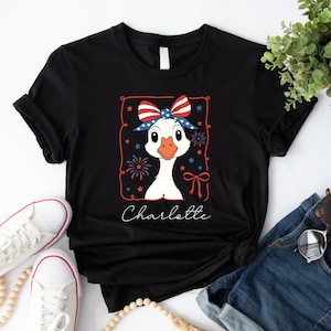 May include: Black t-shirt featuring a cartoon goose wearing a patriotic bow and surrounded by fireworks and stars. The name "Charlotte" is written in cursive below the goose. The shirt is paired with white sneakers and denim.