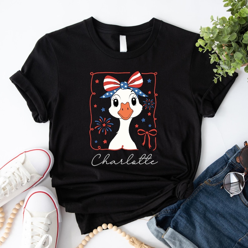 May include: Black t-shirt featuring a cartoon goose wearing a patriotic bow and surrounded by fireworks and stars. The name "Charlotte" is written in cursive below the goose. The shirt is paired with white sneakers and denim.