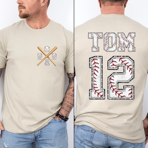 May include: A beige baseball-themed t-shirt. The front features a baseball, crossed bats, and the word "DAD". The back displays "TOM" above the number "12", both with baseball stitching details. The shirt is a casual wear item.