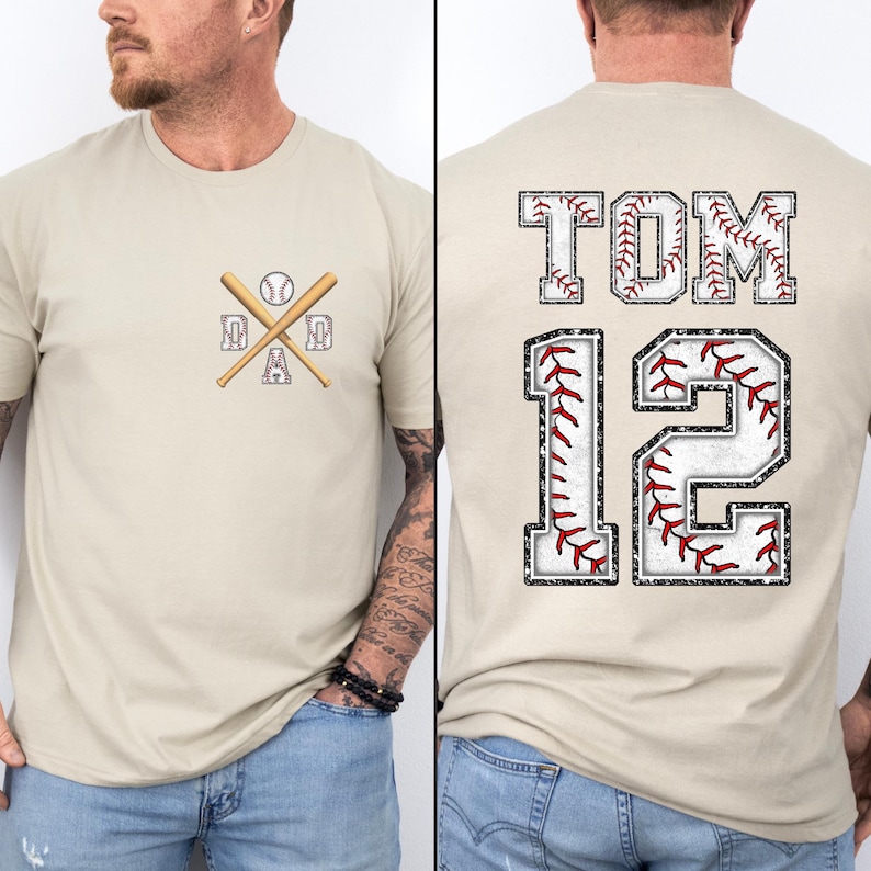 May include: A beige baseball-themed t-shirt. The front features a baseball, crossed bats, and the word "DAD". The back displays "TOM" above the number "12", both with baseball stitching details. The shirt is a casual wear item.