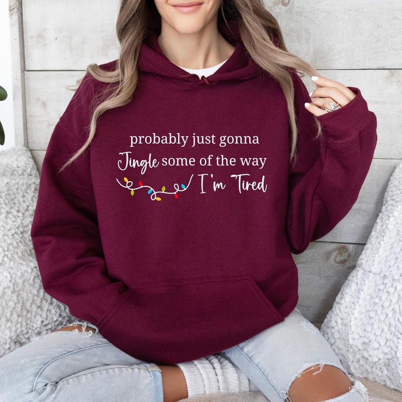 May include: A maroon hooded sweatshirt with the text "probably just gonna Jingle some of the way I'm Tired" in white. A string of colorful Christmas lights is below the text. The sweatshirt has a white undershirt.