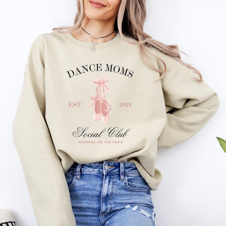May include: A light beige sweatshirt with the text "Dance Moms Est 2024 Social Club Weekends are for Dance" and a pair of pink ballet shoes.