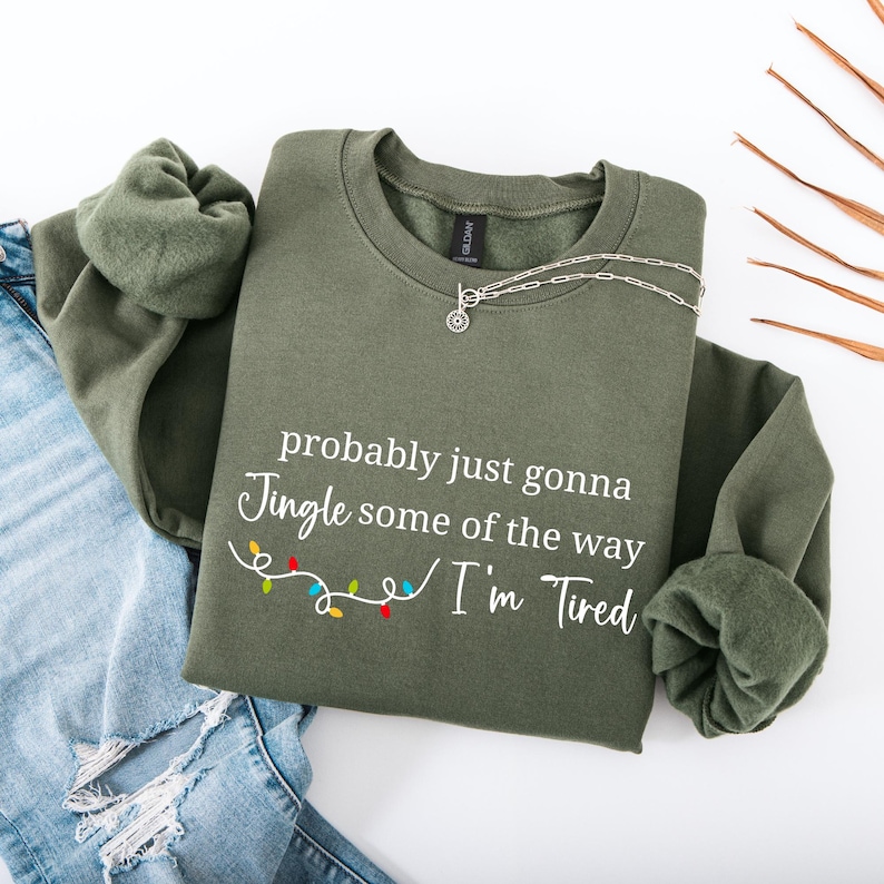 May include: An olive green sweatshirt with the text "probably just gonna Jingle some of the way I'm Tired" in white. A silver necklace with a pendant is on the sweatshirt. The sweatshirt is folded over a pair of blue jeans.