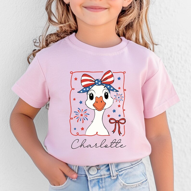 May include: A light pink t-shirt featuring a cartoon goose wearing a patriotic bow and headband. The goose is framed by a red border with stars and fireworks. The name "Charlotte" is written in cursive below the goose.