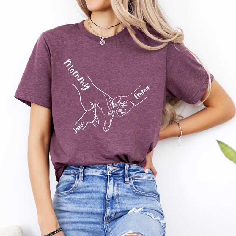 Puede incluir: Una camiseta morada con un contorno blanco de dos manos que se agarran. El texto "Mommy" est&aacute; por encima de las manos, y los nombres "Jane" y "Emma" est&aacute;n por debajo de las manos.