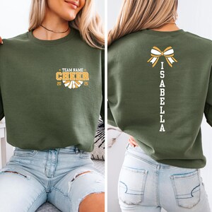 May include: A green sweatshirt with a white bow and the name "ISABELLA" spelled vertically down the back. The front of the sweatshirt has a white and yellow cheerleading pom-pom with the text "TEAM NAME CHEER 20 25".