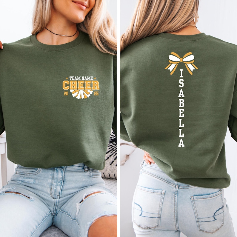 May include: A green sweatshirt with a white bow and the name "ISABELLA" spelled vertically down the back. The front of the sweatshirt has a white and yellow cheerleading pom-pom with the text "TEAM NAME CHEER 20 25".