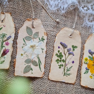 Könnte beinhalten: Vier handgefertigte Papieranhänger mit getrockneten Blumen und Blättern. Jeder Anhänger zeigt eine einzigartige Anordnung von gepressten Blumen in Lila-, Weiß- und Gelbtönen. Die Anhänger haben einen rustikalen, zerrissenen Rand und sind mit Bindfaden aufgefädelt.