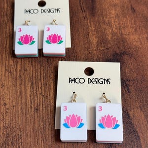 Puede incluir: Dos pares de pendientes colgantes con fichas de mahjong rectangulares blancas, un gr&aacute;fico de flor de loto rosa y el n&uacute;mero 3 en rosa. Cada par est&aacute; sujeto a una tarjeta de presentaci&oacute;n color crema con el texto PACO DESIGNS impreso en negro.