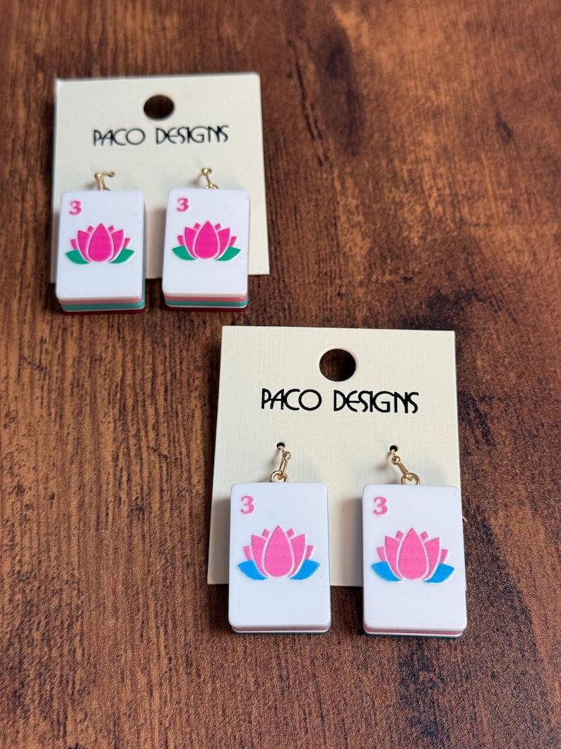 Puede incluir: Dos pares de pendientes colgantes con fichas de mahjong rectangulares blancas, un gr&aacute;fico de flor de loto rosa y el n&uacute;mero 3 en rosa. Cada par est&aacute; sujeto a una tarjeta de presentaci&oacute;n color crema con el texto PACO DESIGNS impreso en negro.