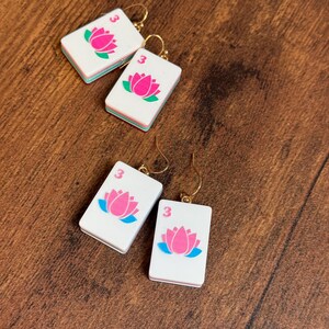Puede incluir: Dos pares de pendientes rectangulares blancos con forma de fichas de mahjong, cada uno con un gr&aacute;fico de flor de loto rosa, un peque&ntilde;o n&uacute;mero tres rojo y ganchos de metal dorado. Las fichas tienen un dise&ntilde;o de borde en capas blanco, verde y blanco.