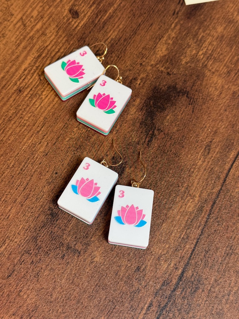 Puede incluir: Dos pares de pendientes rectangulares blancos con forma de fichas de mahjong, cada uno con un gr&aacute;fico de flor de loto rosa, un peque&ntilde;o n&uacute;mero tres rojo y ganchos de metal dorado. Las fichas tienen un dise&ntilde;o de borde en capas blanco, verde y blanco.
