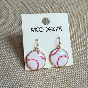 May include: Baseball-themed earrings with a white background and red stitching, set in a gold-colored circular frame. The earrings are displayed on a white card with the text "PACO DESIGNS".