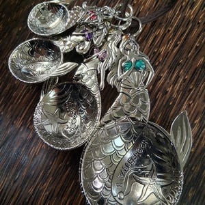 May include: A set of silver measuring spoons with mermaid designs. Each spoon is shaped like a mermaid with a detailed tail and a starfish accent. The spoons are connected by a brown leather cord. Measurements include 1 tablespoon, 1 teaspoon, and 1/2 teaspoon.