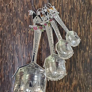 May include: A set of silver measuring spoons with decorative cow and barn designs. Each spoon is labeled with a measurement, including tablespoon, teaspoon, and fractions thereof. The spoons are attached to a brown leather loop for hanging.