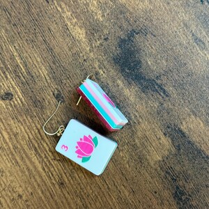 Puede incluir: Un par de pendientes rectangulares con una superficie blanca, un gr&aacute;fico de flor de loto rosa y el n&uacute;mero 3 en rosa. Los pendientes tienen un perfil lateral en capas con secciones en blanco, turquesa y rosa oscuro y ganchos de metal dorado.