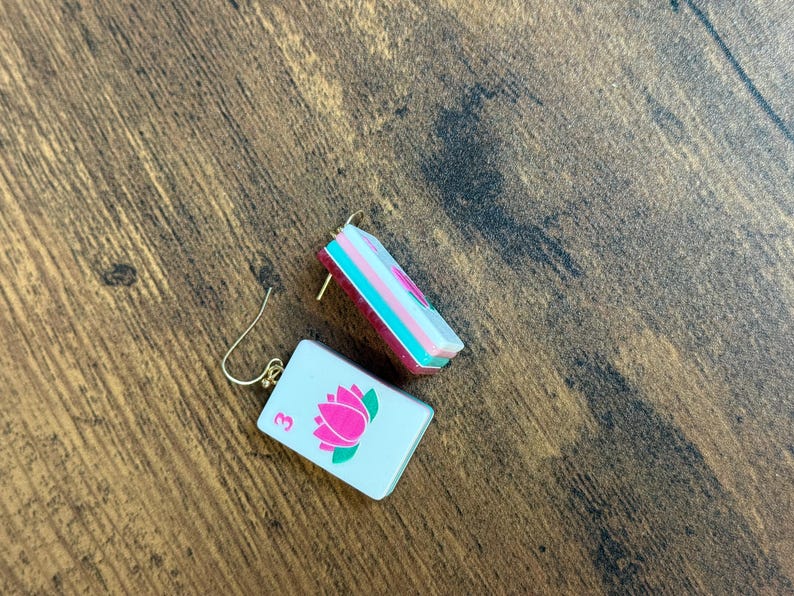 Puede incluir: Un par de pendientes rectangulares con una superficie blanca, un gr&aacute;fico de flor de loto rosa y el n&uacute;mero 3 en rosa. Los pendientes tienen un perfil lateral en capas con secciones en blanco, turquesa y rosa oscuro y ganchos de metal dorado.